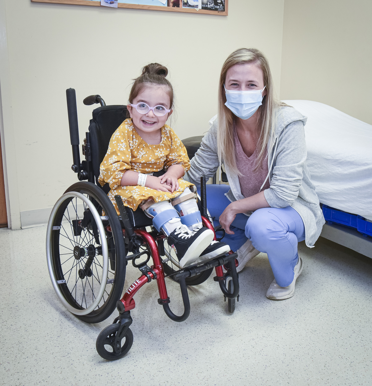 Young female Shriners patient in wheelchair smiles alongside nurse in hospital